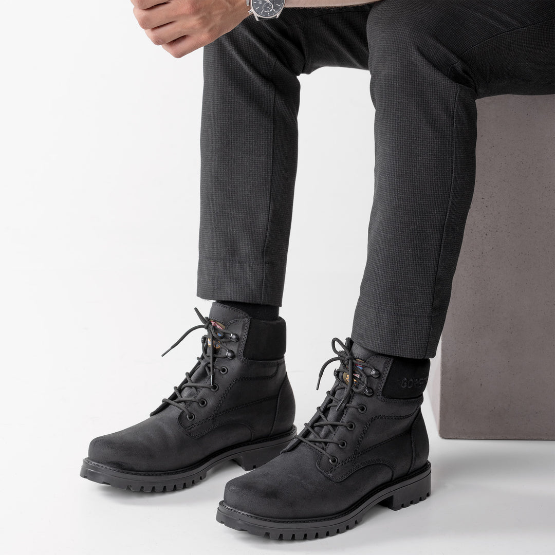 Kuva miesten mustasta talvikengästä henkilön jalassa valkoisella taustalla.