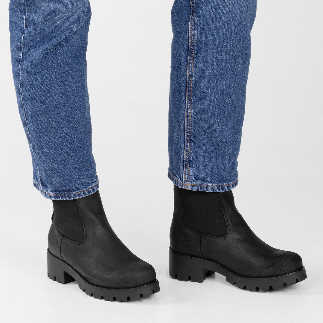 Malli seisoo valkoisella taustalla jalassaan vahanupukkinahkaiset mustat naisten Chelsea-nilkkurit.