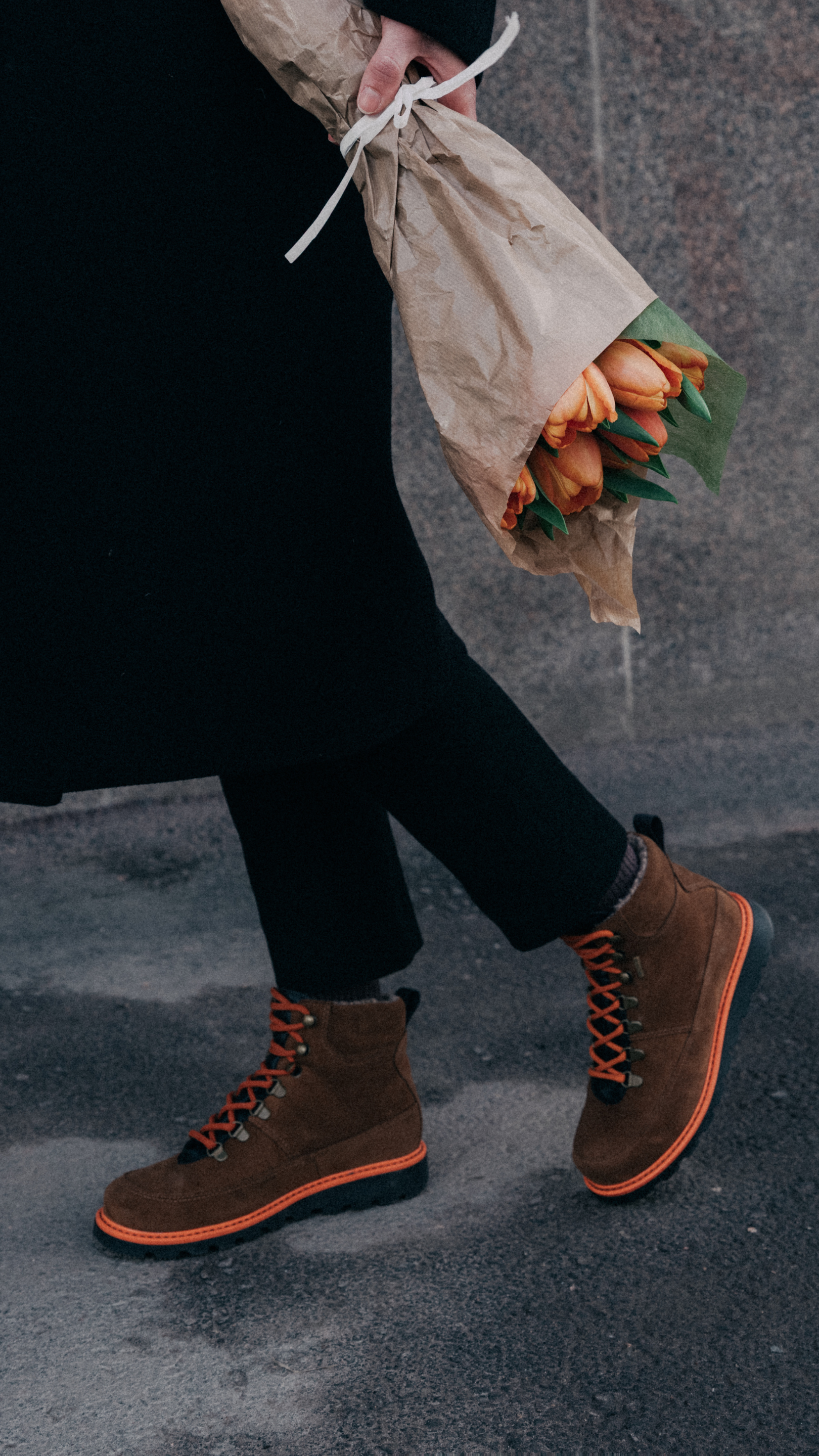 Henkilö kävelee ulkona jalassaan kengät oransseilla yksityiskohdilla ja kukkia kädessään.