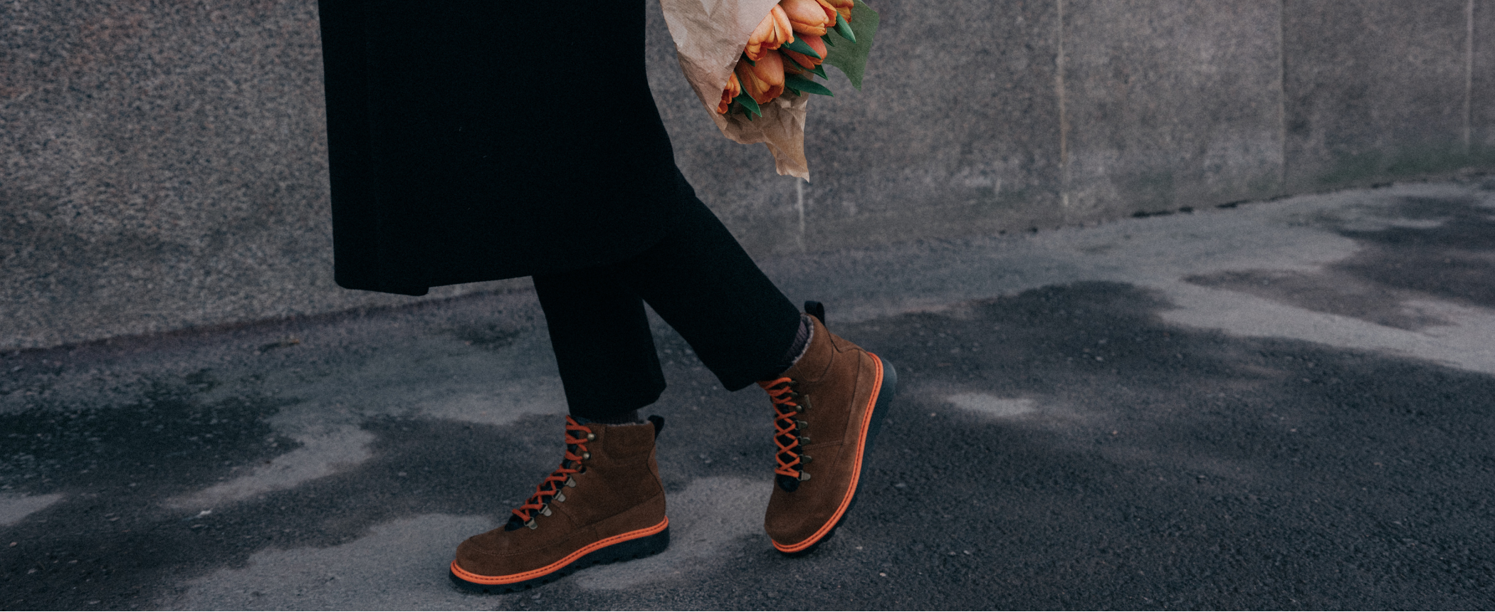 Henkilö kävelee ulkona jalassaan oransseilla yksityiskohdilla varustetut kengät, kädessään oranssit kukat.
